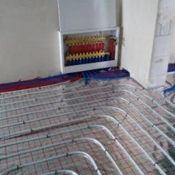 Rozłożone rury ogrzewania podłogowego na folii z siatką, widoczna szafka rozdzielacza z czerwonymi i niebieskimi rurami.