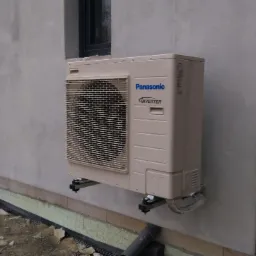 Zewnętrzna jednostka klimatyzacji Panasonic Inverter zamontowana na ścianie budynku z szarą elewacją i czarnymi drzwiami, widoczne rury instalacyjne i elementy montażowe.