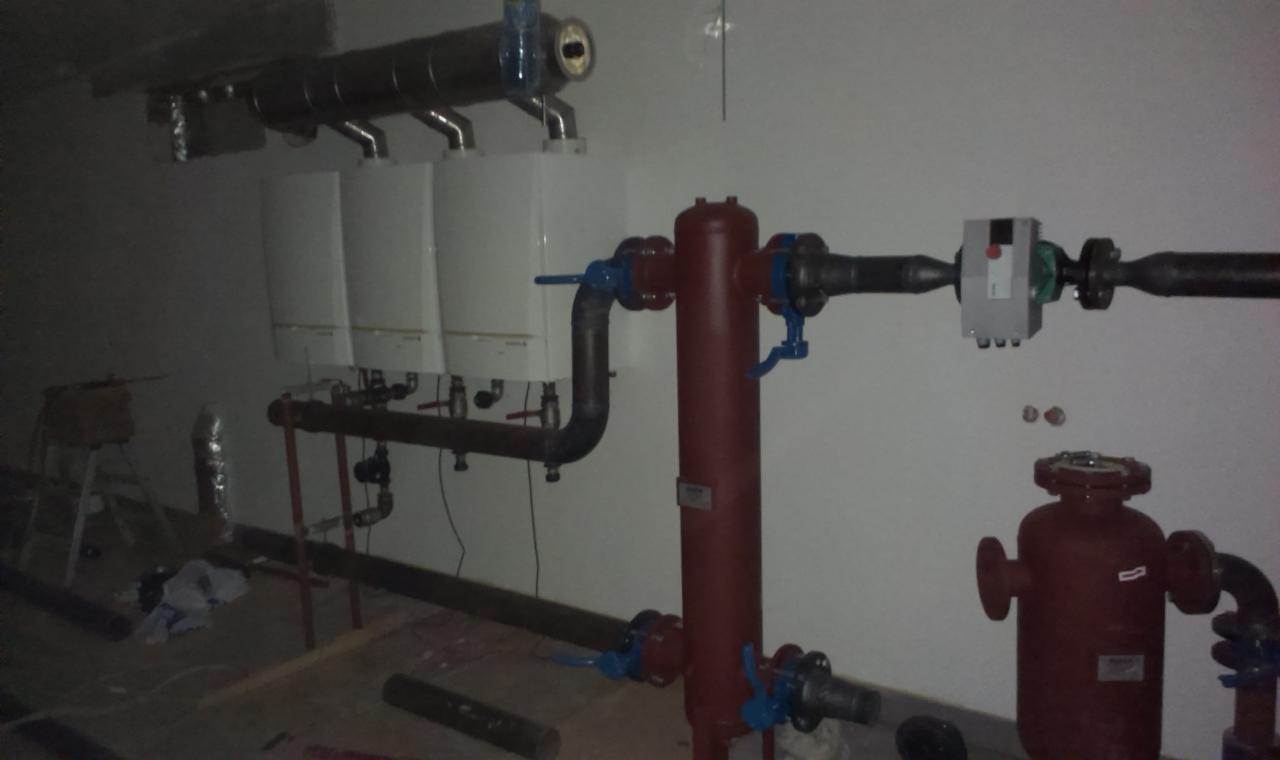 Instalacja kotłów grzewczych z rurami i zaworami, widoczne elementy instalacji hydraulicznej, w tle drabina i nieporządek na podłodze.
