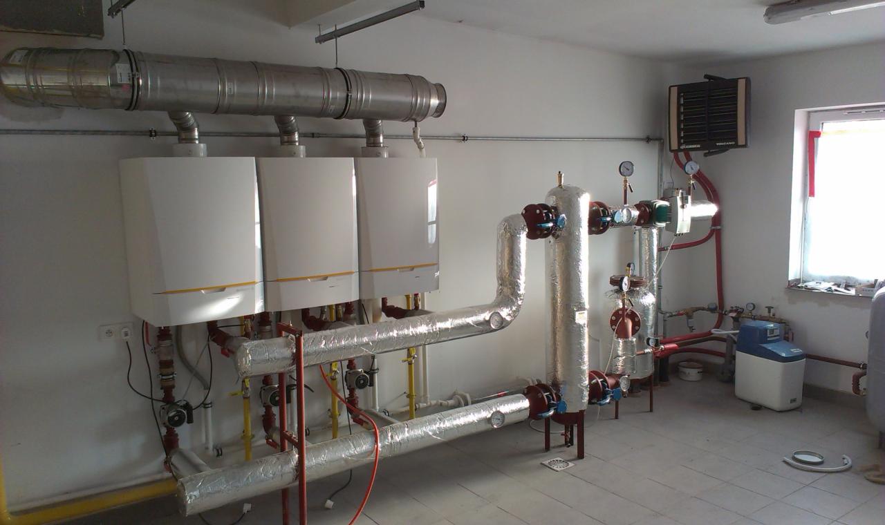 Instalacja trzech białych kotłów grzewczych z żółtymi akcentami, połączonych rurami w srebrnej izolacji, z dodatkowymi elementami instalacji hydrauliczej, w jasnym pomieszczeniu z płytkami...
