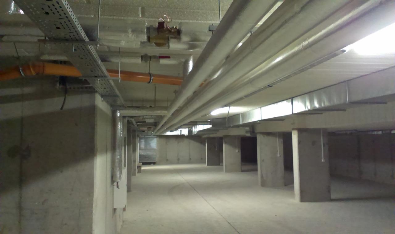 Instalacja wentylacyjna i rury pod sufitem w surowym, betonowym pomieszczeniu, widoczne koryta kablowe i zawór z czerwonym pokrętłem.