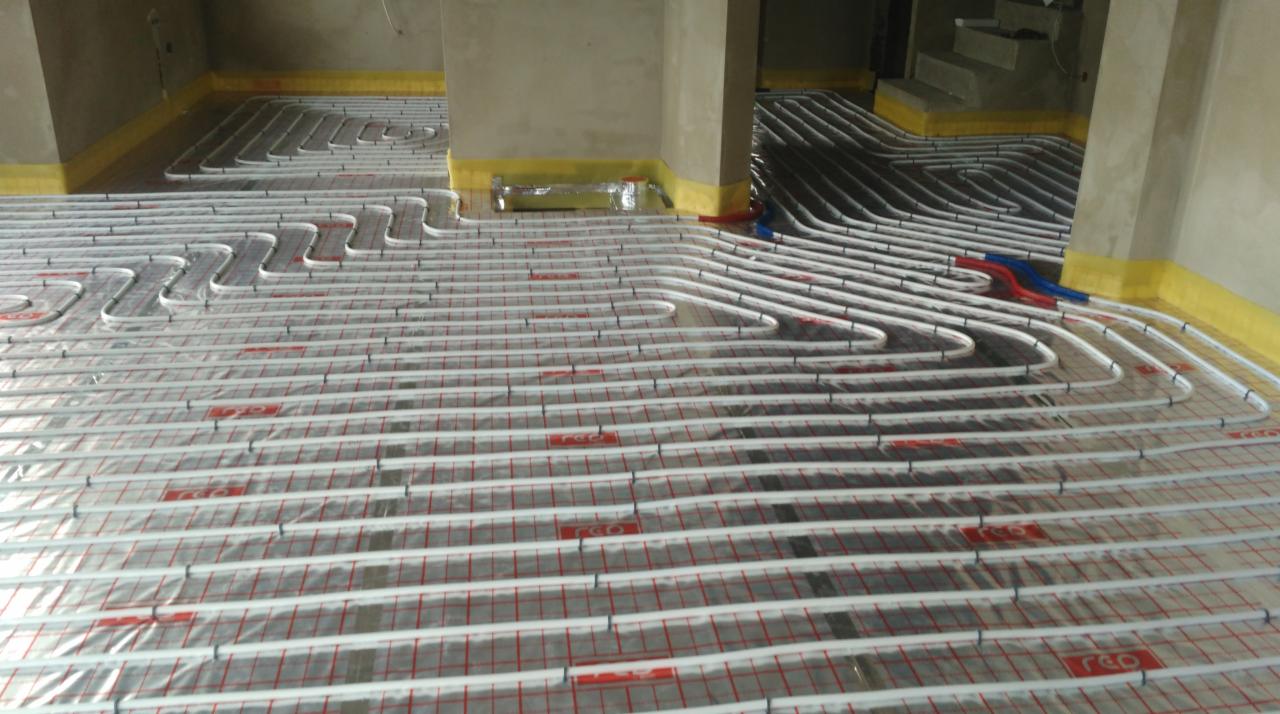 Rozłożona instalacja ogrzewania podłogowego w surowym pomieszczeniu, rury grzewcze zamocowane na folii z siatką, widoczne fragmenty ścian zabezpieczone taśmą ochronną, w tle schody w stanie surowym.