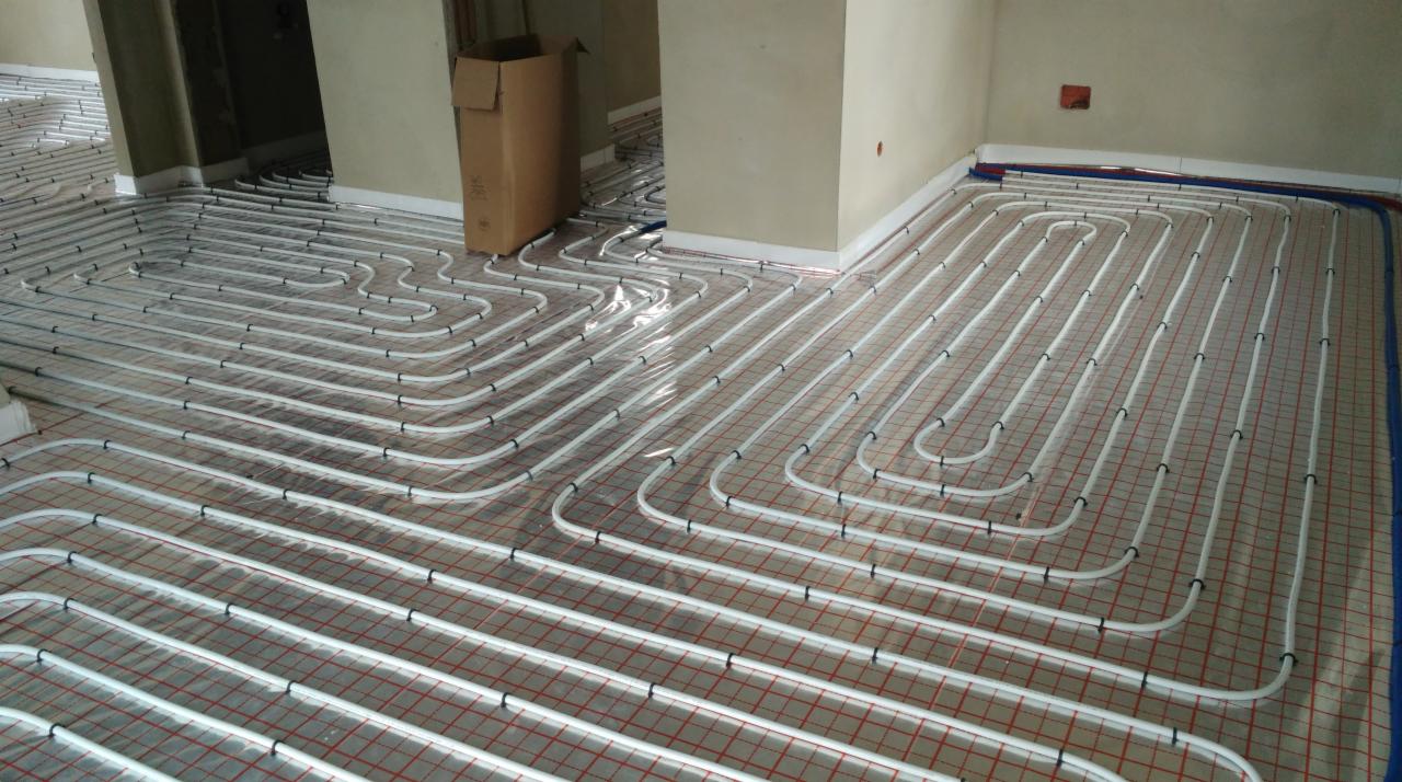 Białe rury ogrzewania podłogowego ułożone na czerwonej siatce, widoczne w kilku pomieszczeniach w trakcie instalacji, z kartonem w tle.