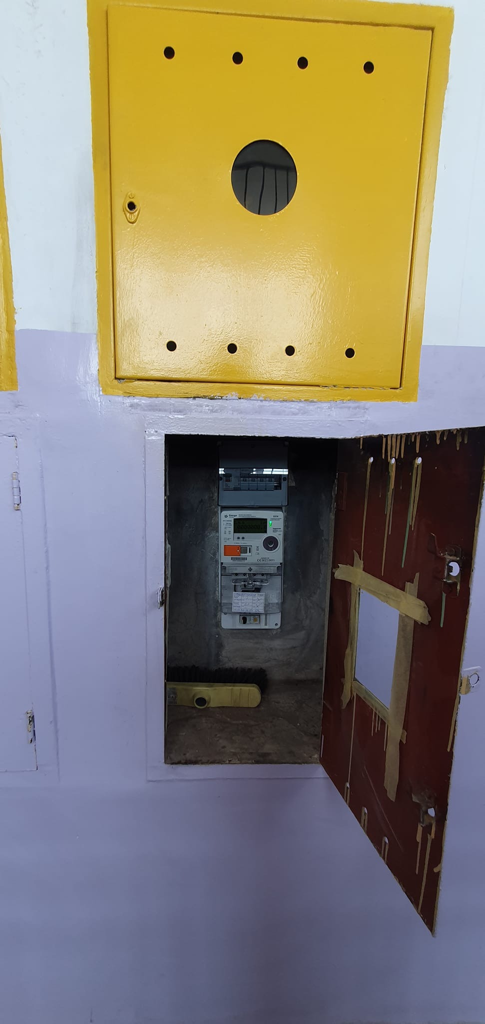 Otwarta skrzynka z licznikiem elektrycznym wewnątrz, widoczna poziomica i ślady po malowaniu na ścianie i drzwiach.