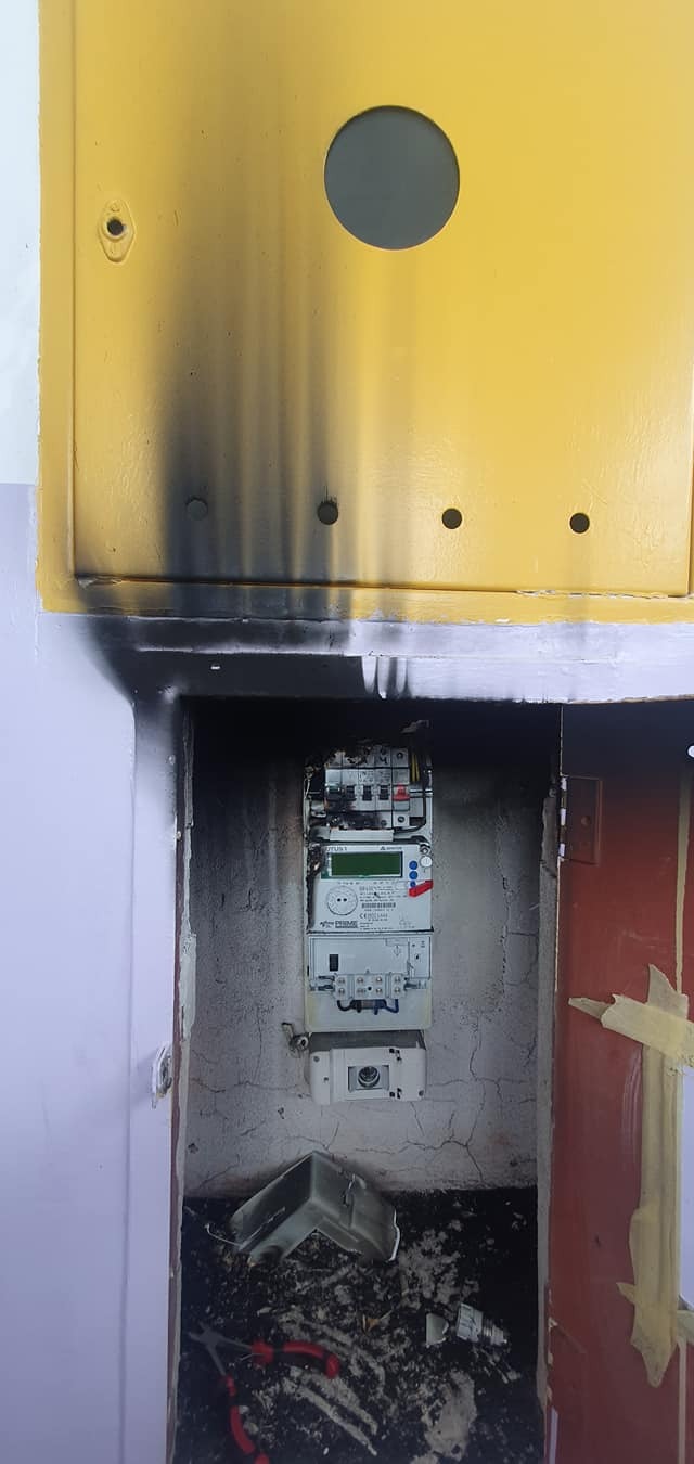 Uszkodzona skrzynka z licznikiem elektrycznym, widoczne ślady okopcenia, resztki plastiku na podłodze, szczypce leżące obok.