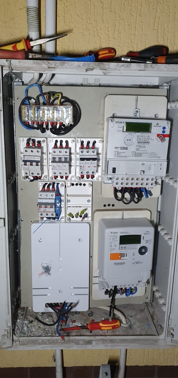Otwarta skrzynka elektryczna z zamontowanymi licznikami, bezpiecznikami i przewodami, widoczne narzędzia na górnej półce i w dolnej części skrzynki.