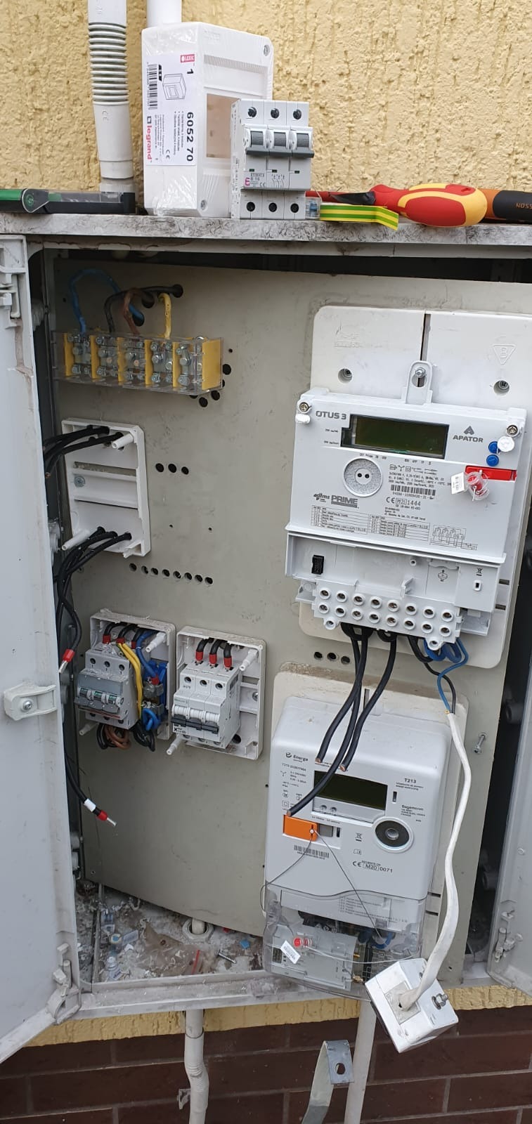 Otwarta skrzynka elektryczna z zamontowanymi licznikami energii, zabezpieczeniami i złączkami, widoczne kable, narzędzia i elementy montażowe na górnej półce, w tle fragment elewacji budynku.