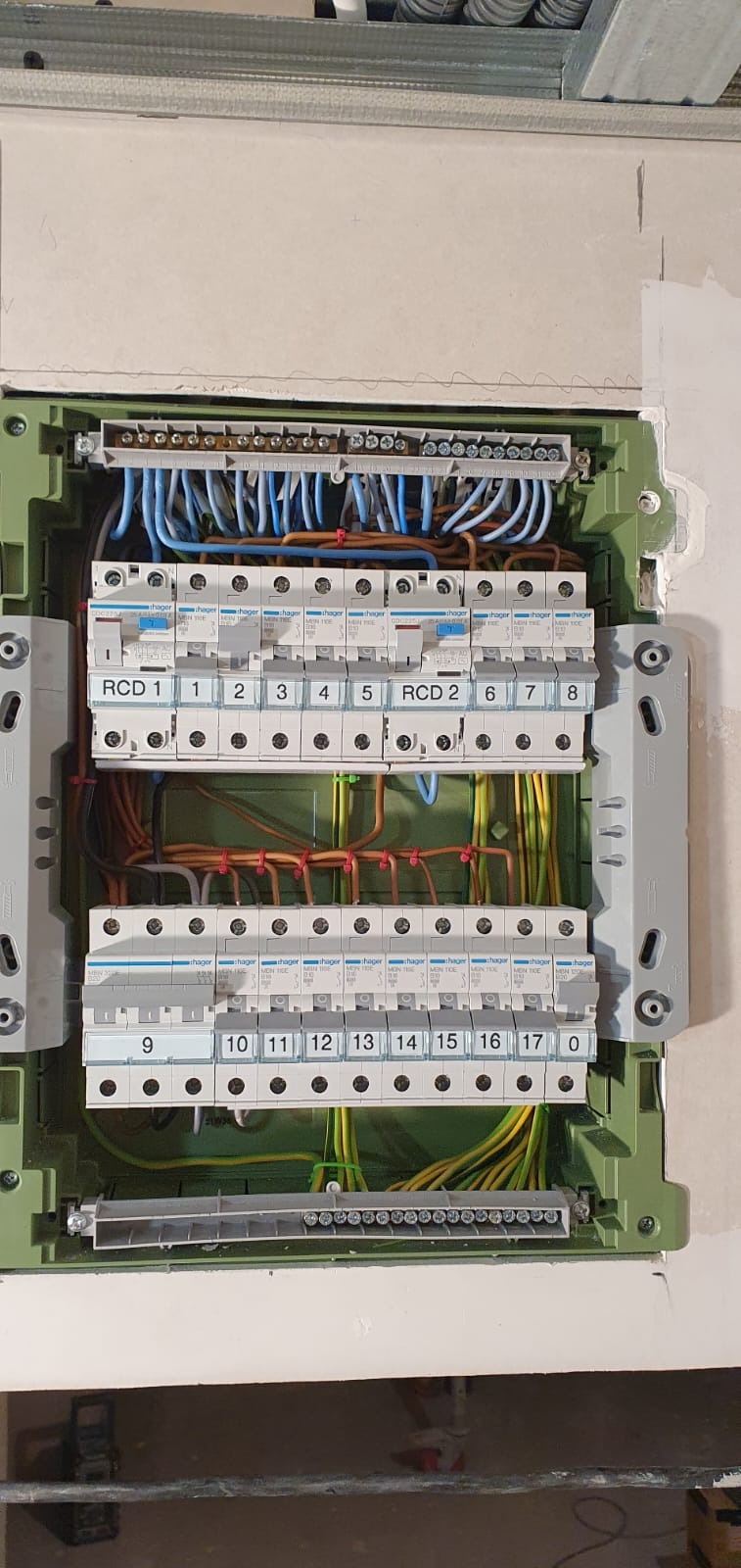 Otwarta skrzynka elektryczna z zainstalowanymi bezpiecznikami Hager i podłączonymi przewodami w kolorach niebieskim, brązowym, zielono-żółtym i czarnym, widok z przodu, ściana w tle przygotowana...
