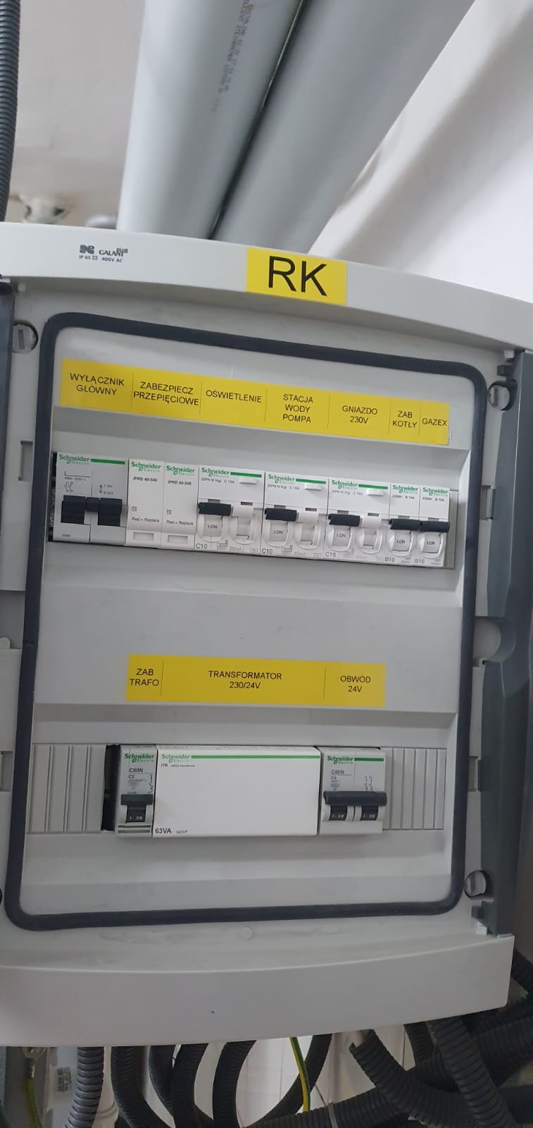 Rozdzielnica elektryczna Schneider z wyłącznikami i zabezpieczeniami, oznaczone żółtymi etykietami informacyjnymi, widoczne przewody w peszlach.