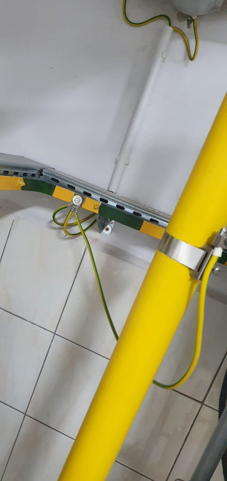 Żółta rura instalacyjna przymocowana metalową obejmą, widoczne uziemienie z kablem żółto-zielonym oraz metalowa szyna montażowa z elementami mocującymi na tle białej ściany i kafelków.