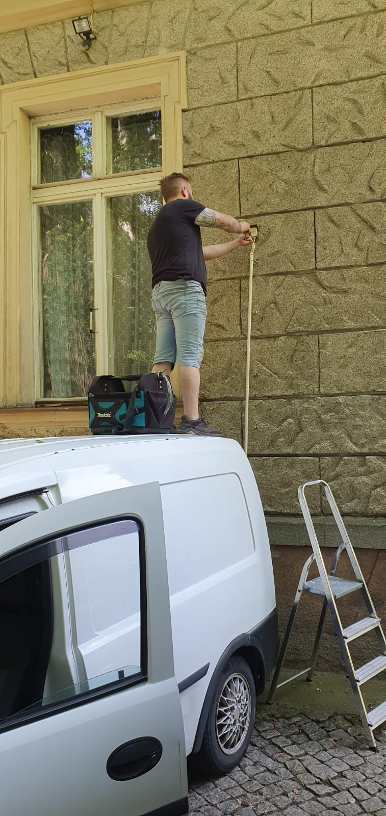 Elektryk z tatuażami na ramionach, stojący na dachu białego furgonetki z otwartymi drzwiami, przy zabytkowej elewacji budynku, instaluje czujnik zmierzchowy; obok stoi drabina.