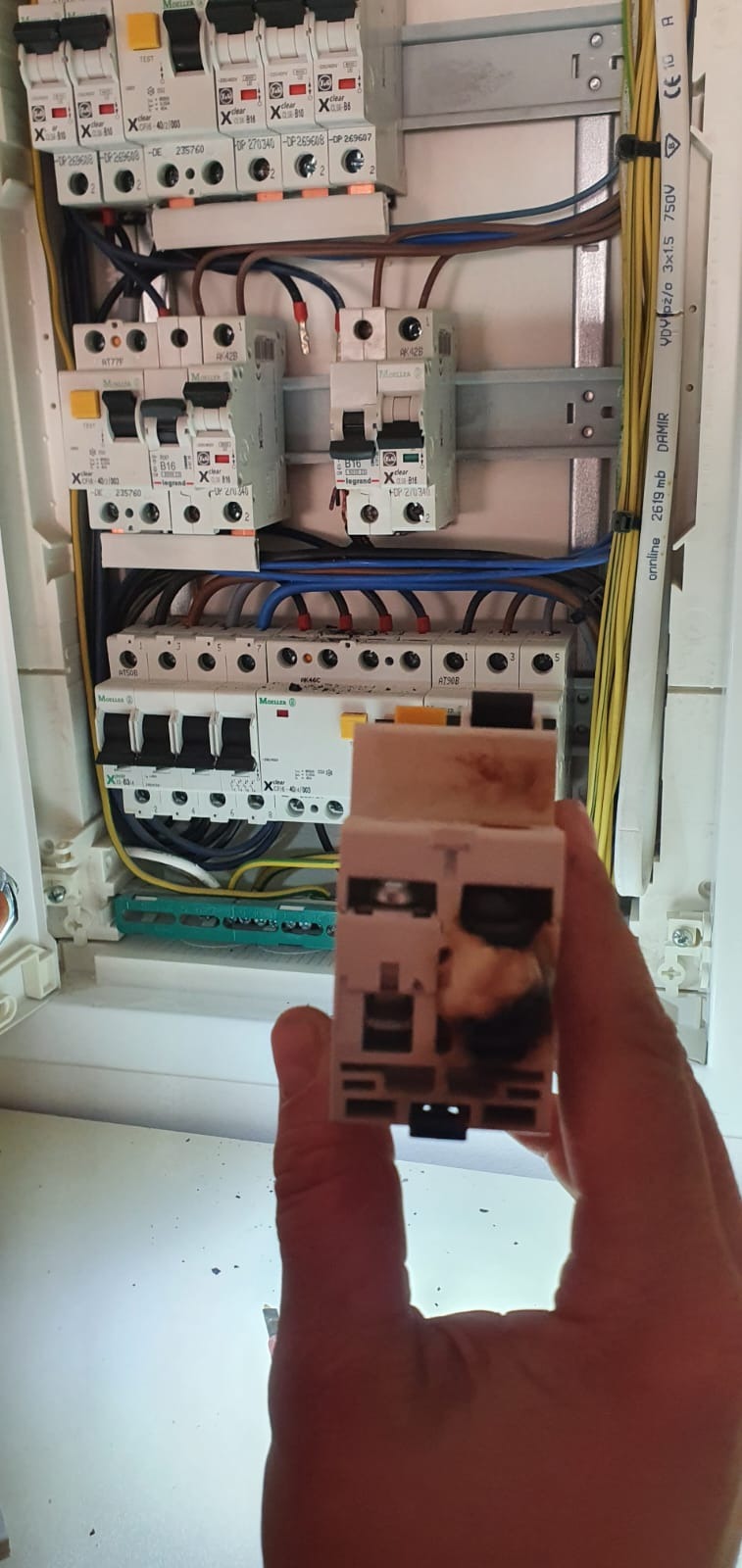 Uszkodzony wyłącznik różnicowoprądowy w dłoni na tle skrzynki z bezpiecznikami, widoczne ślady przepalenia na obudowie.