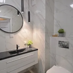 Nowoczesna łazienka z marmurowymi płytkami, wiszącą szafką z czarnym blatem, okrągłym lustrem i minimalistycznym oświetleniem punktowym.