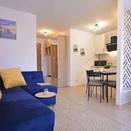 Nowoczesne wnętrze mieszkania z granatowym narożnikiem, okrągłym stolikiem, aneksem kuchennym i dekoracyjną ścianką działową w geometryczny wzór.