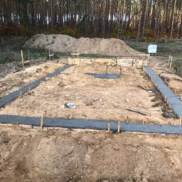 Wylane fundamenty pod budynek na działce z widokiem na las, szalunki z drewnianych kołków, w tle hałda ziemi i biały zbiornik.