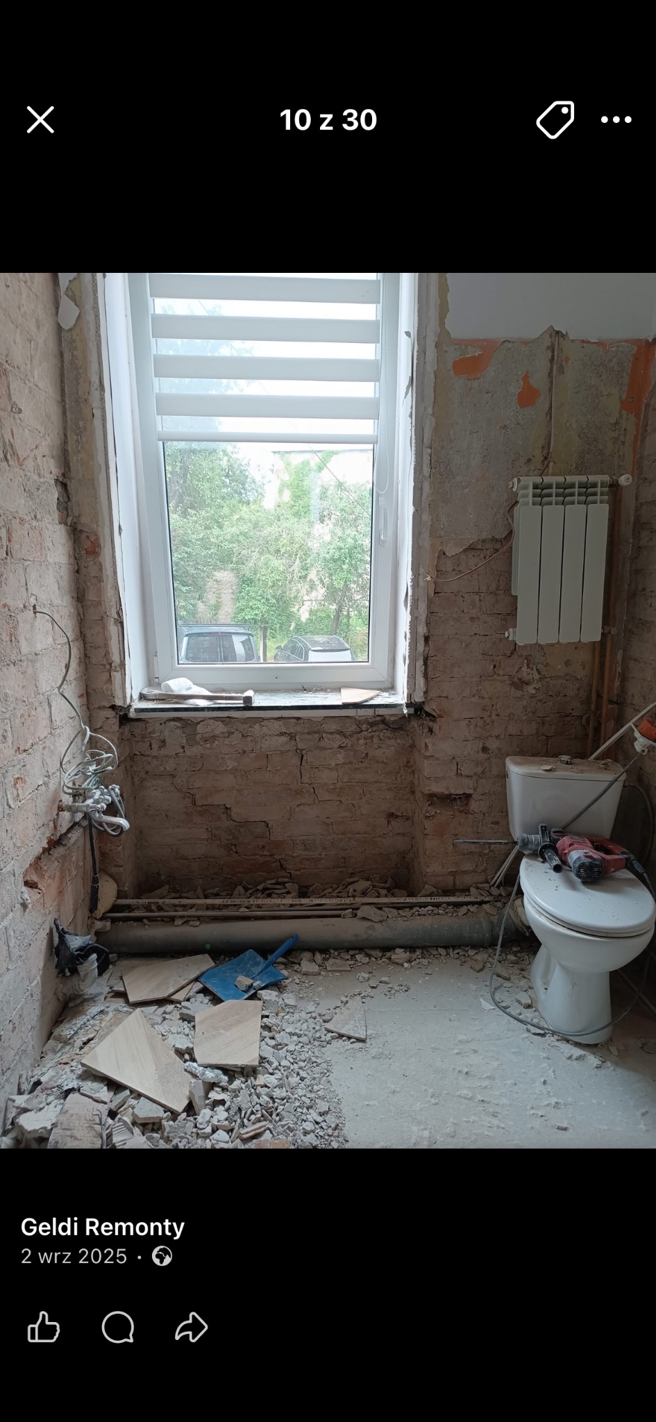 Łazienka w trakcie demontażu: odsłonięta cegła, gruz, narzędzia na sedesie. Widoczne rury, okno z żaluzjami i grzejnik. Etap przygotowawczy do remontu.