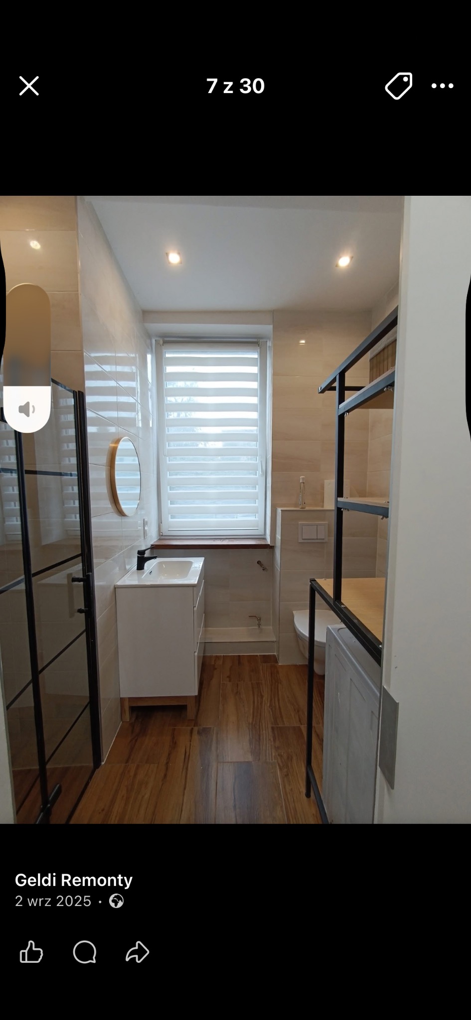 Jasna łazienka po remoncie z drewnopodobną podłogą, kabiną prysznicową z czarnymi ramami i białą szafką z umywalką. Widoczne okno z żaluzjami i metalowy regał.