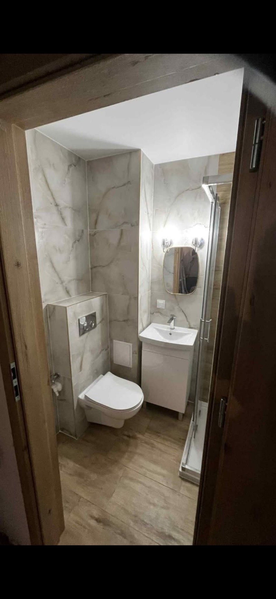 Wykończona łazienka z kabiną prysznicową, umywalką z szafką i podwieszaną toaletą. Ściany i podłoga wyłożone płytkami imitującymi marmur i drewno.
