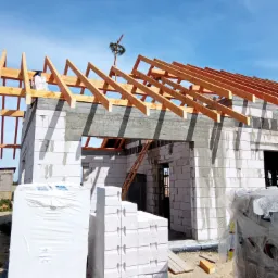 Dom murowany w trakcie budowy, widoczne białe bloczki, drewniana konstrukcja dachu z wieńcem na szczycie, materiały budowlane na pierwszym planie.