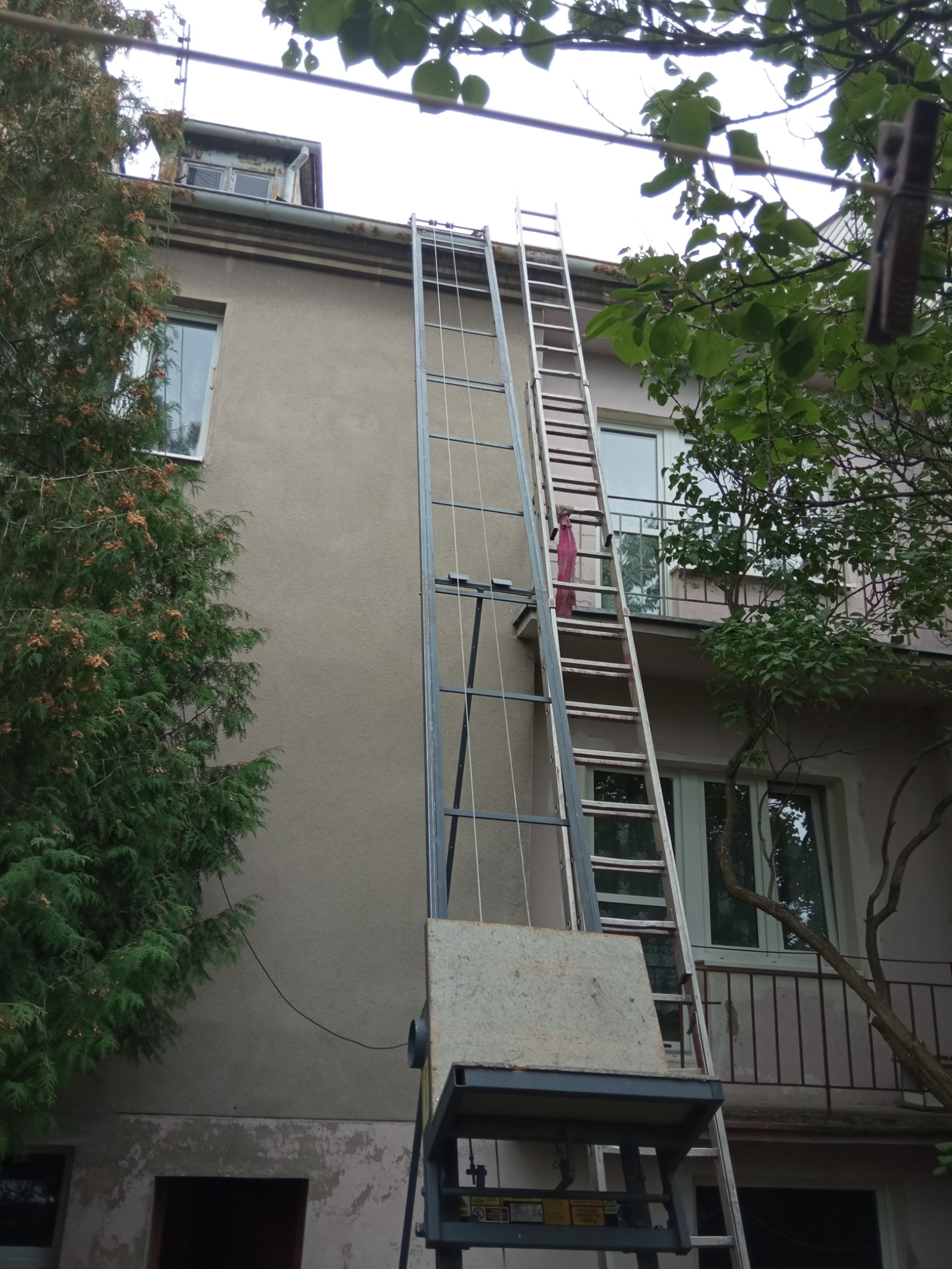 Platforma podnośnikowa ustawiona przy ścianie budynku mieszkalnego, obok drabina oparta o balkon. Widoczne drzewa i fragment dachu.
