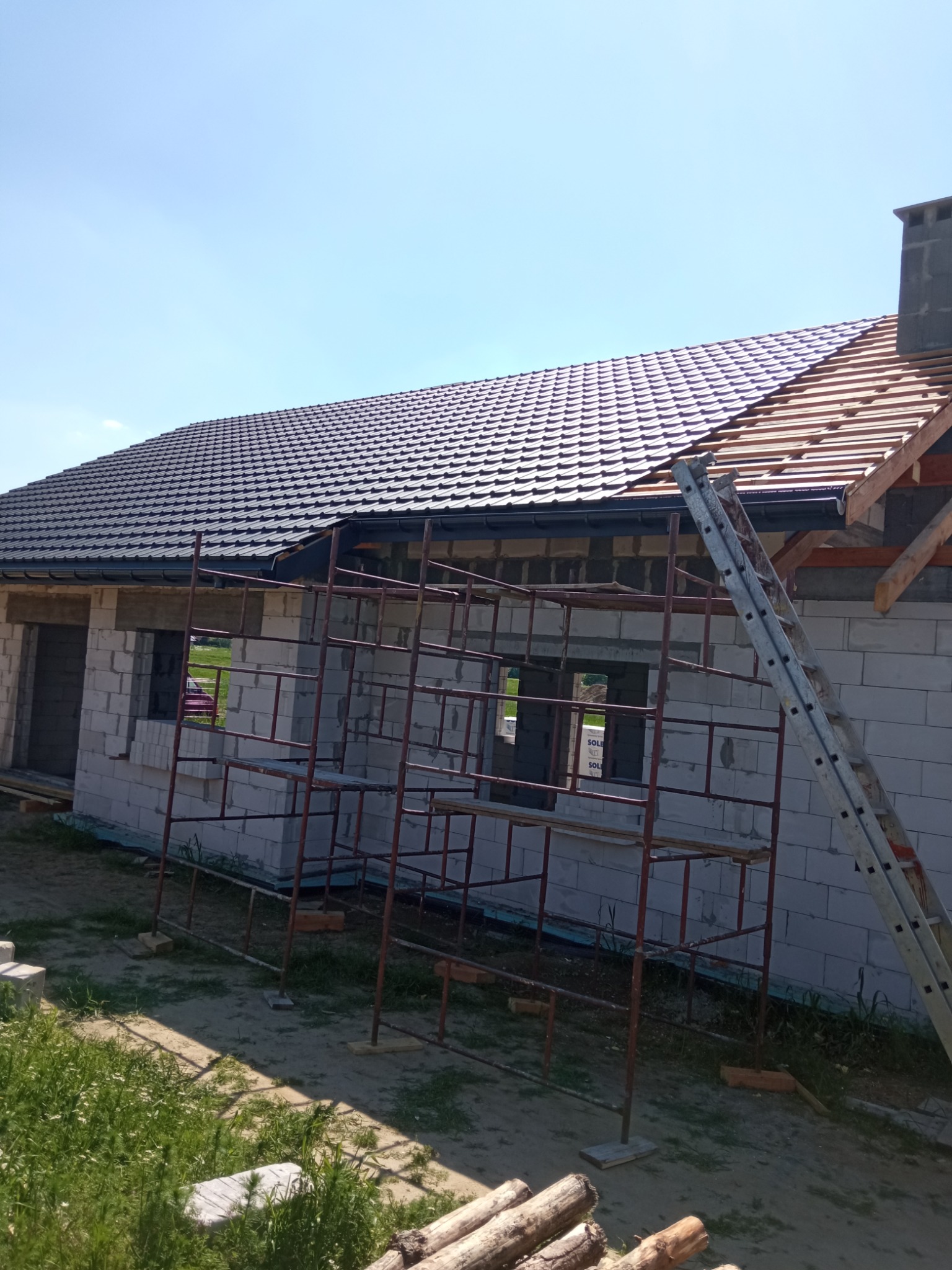Nowo budowany dom z białego pustaka z częściowo ukończonym dachem z ciemnej dachówki ceramicznej, widoczne rusztowania i drabina oparta o budynek.