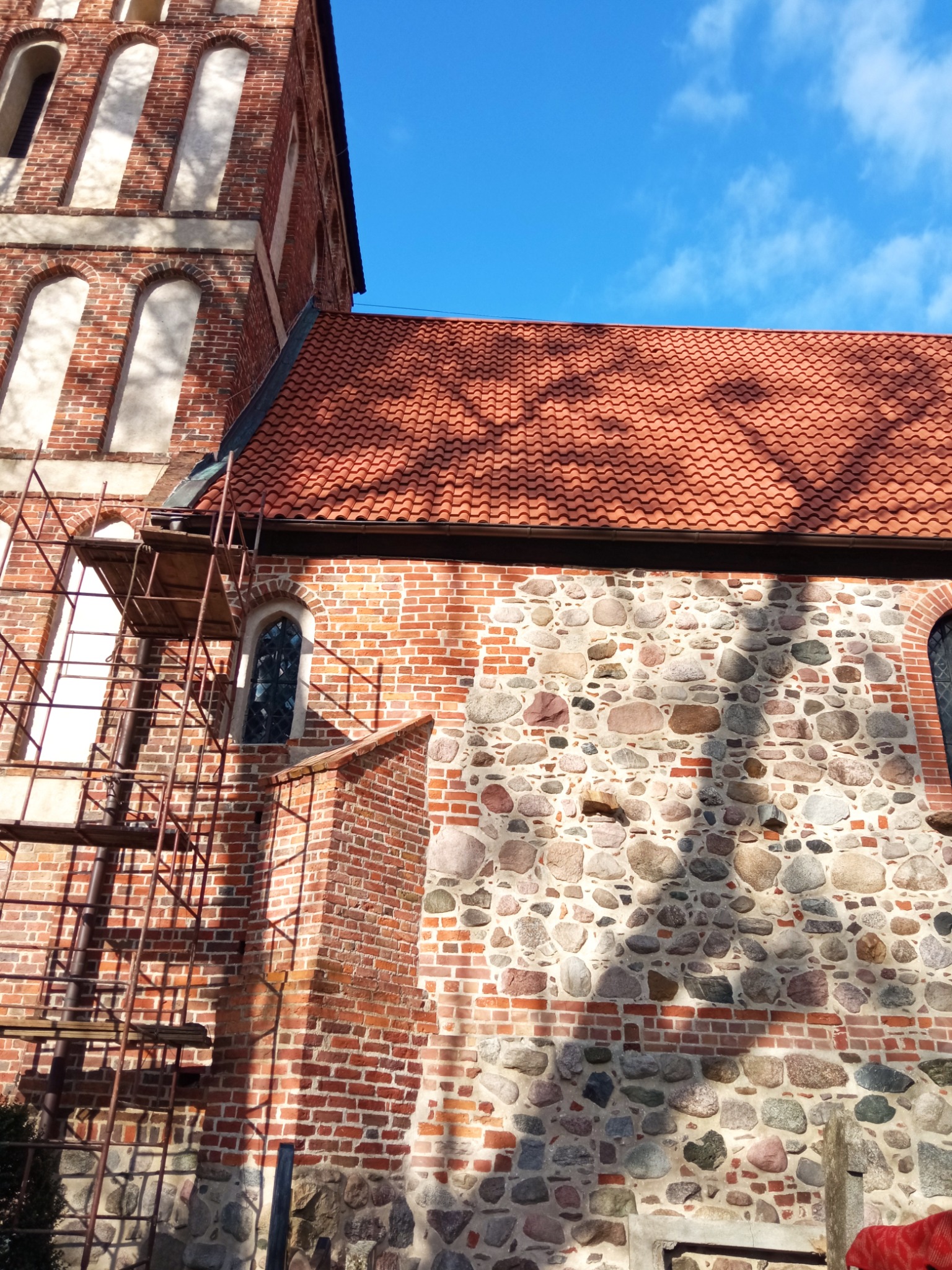 Ceglano-kamienna wieża kościoła z nową, czerwoną dachówką, częściowo zasłonięta rusztowaniem w słoneczny dzień.