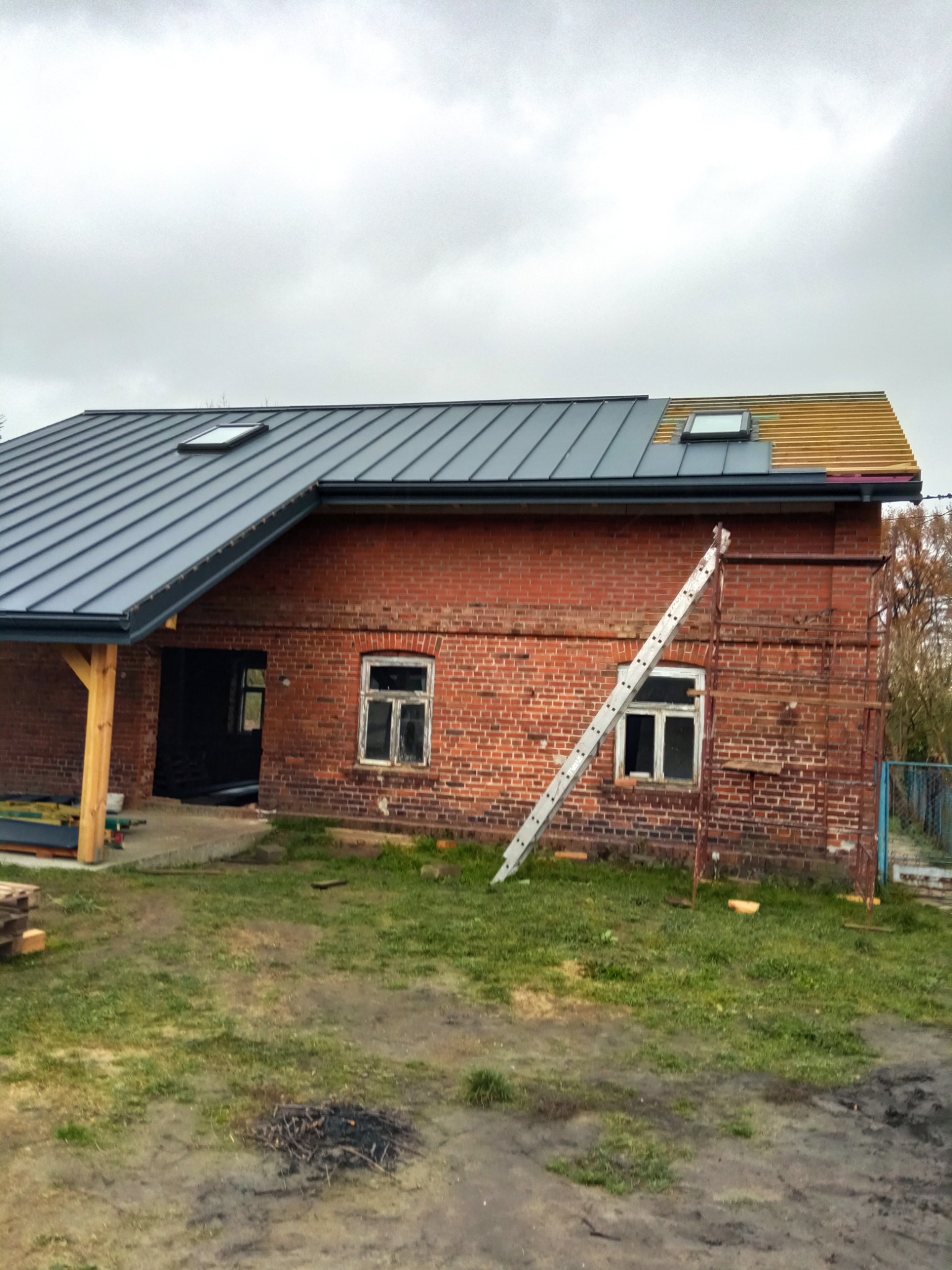 Częściowo wykończony dach z blachy na murowanym domu z czerwonej cegły, z oknami dachowymi i drabiną opartą o ścianę, widok na budynek z zewnątrz w pochmurny dzień.