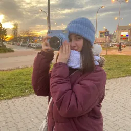 Młoda kobieta w zimowej odzieży robi zdjęcie aparatem Instax na tle zachodzącego słońca i zabudowań miejskich.