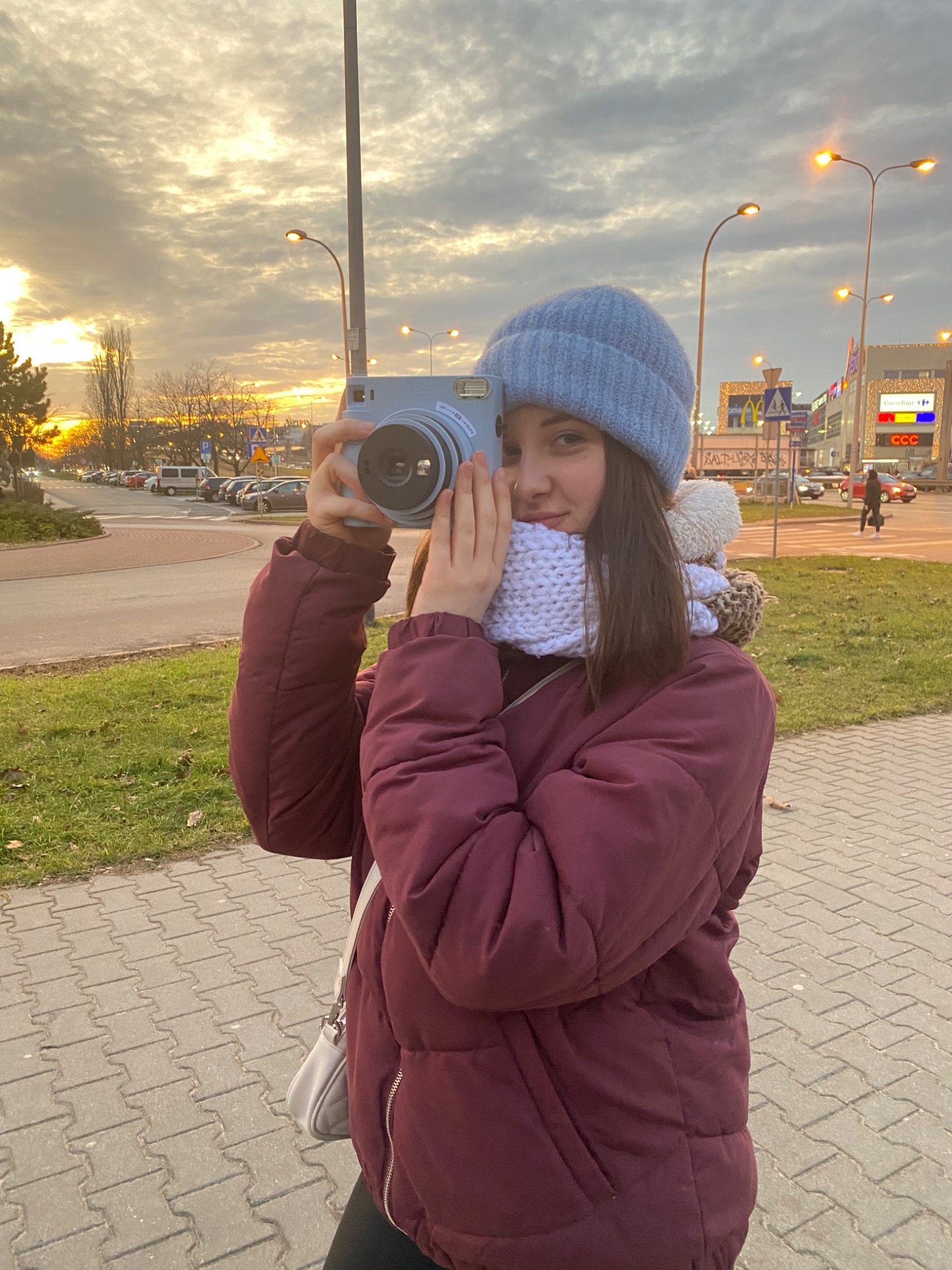 Młoda kobieta w zimowej odzieży robi zdjęcie aparatem Instax na tle zachodzącego słońca i zabudowań miejskich.