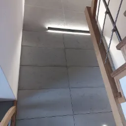 Ściana wykończona płytami imitującymi beton z oświetleniem punktowym i liniowym, fragment drewnianych schodów z metalową balustradą po prawej stronie.