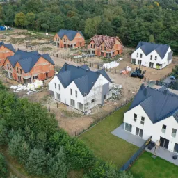 Widok z lotu ptaka na osiedle domów jednorodzinnych w różnym stadium budowy, od surowej cegły po domy z panelami słonecznymi i zagospodarowanym terenem.