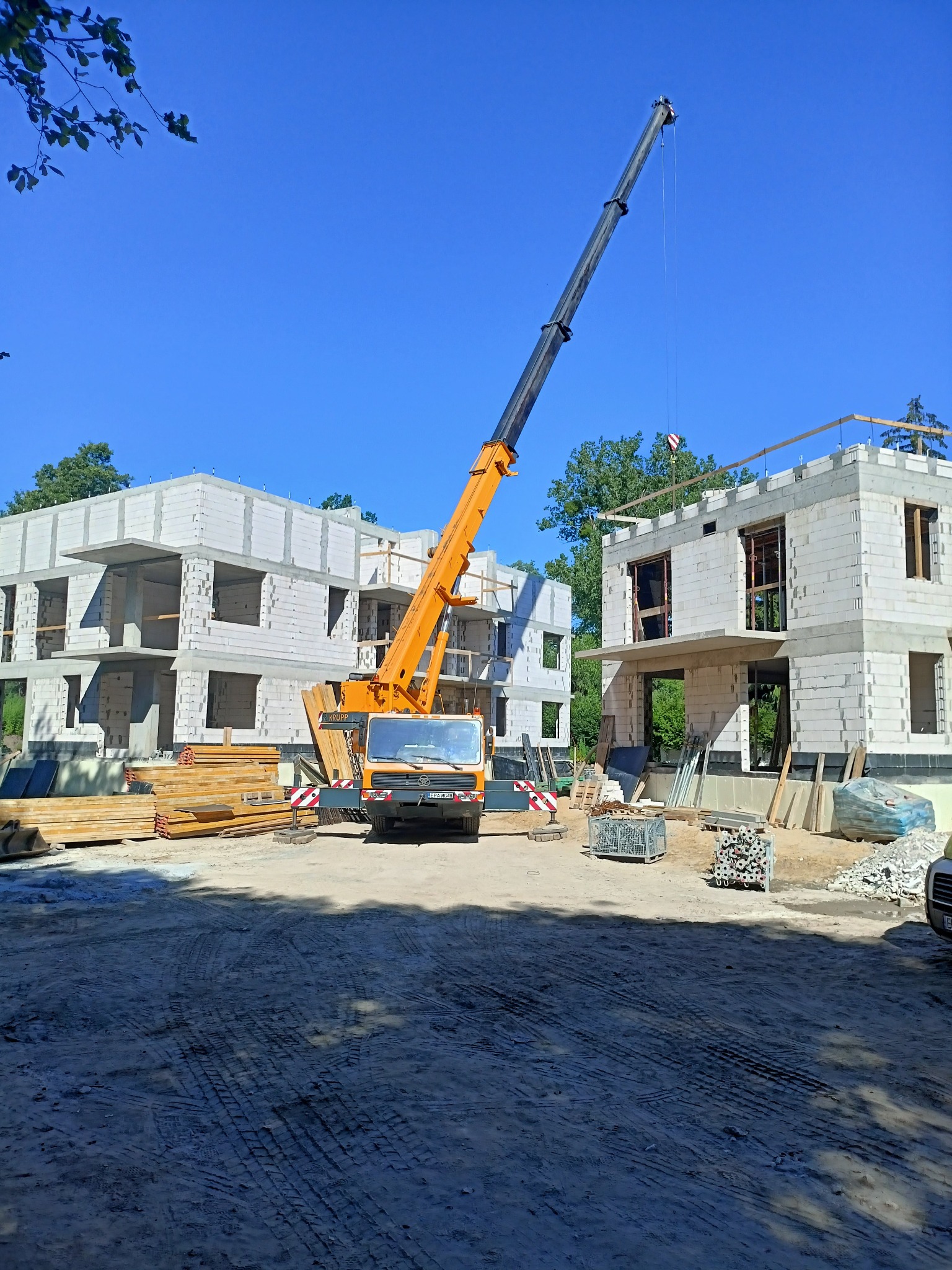 Żuraw budowlany na placu budowy, w tle dwa wznoszone budynki z białych bloczków, widoczne drewniane elementy konstrukcyjne.
