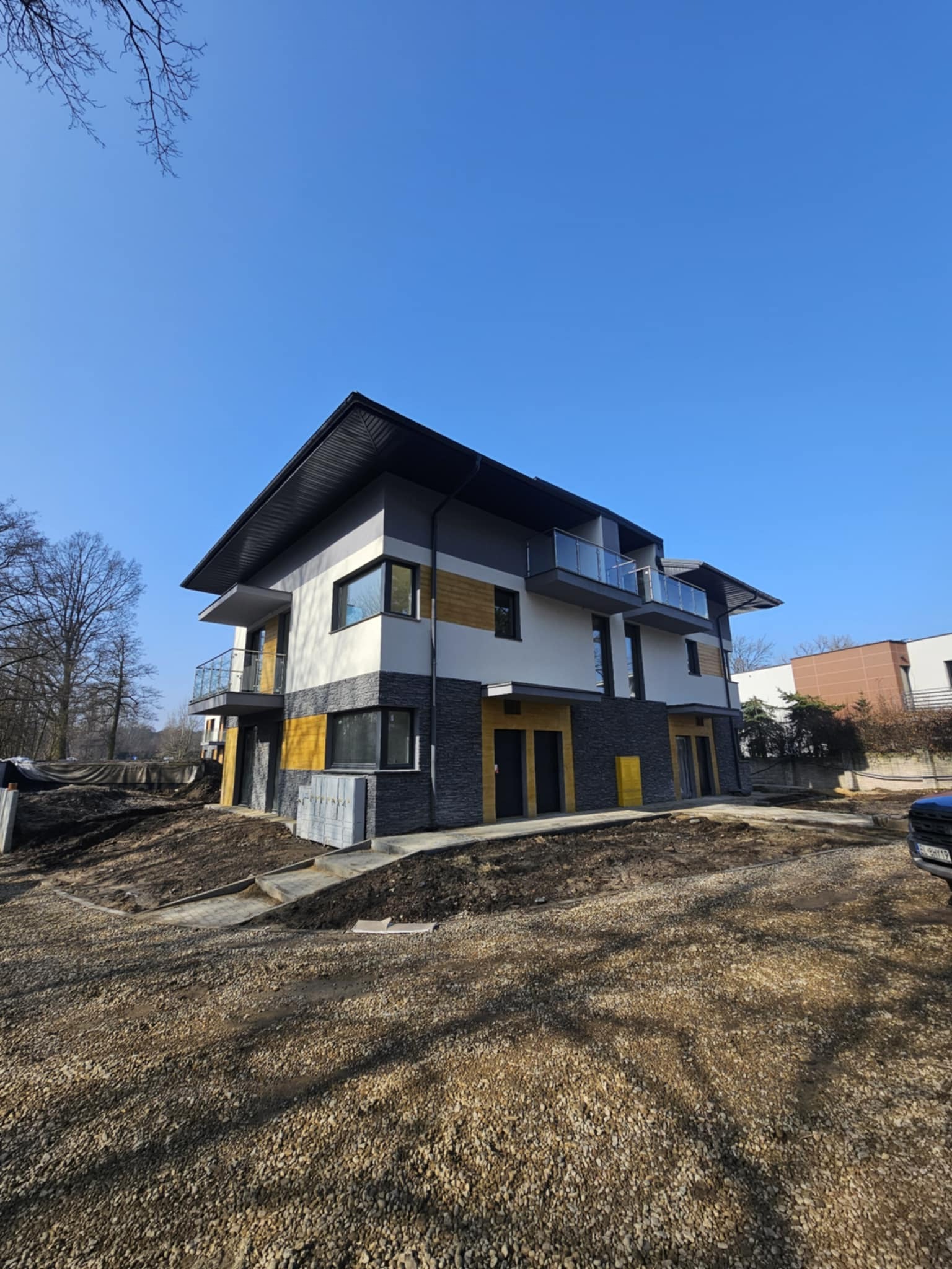 Dwukondygnacyjny dom z płaskim dachem, wykończony w kolorach szarości, bieli i żółci, z balkonami ze szklanymi balustradami, na działce w trakcie zagospodarowywania.