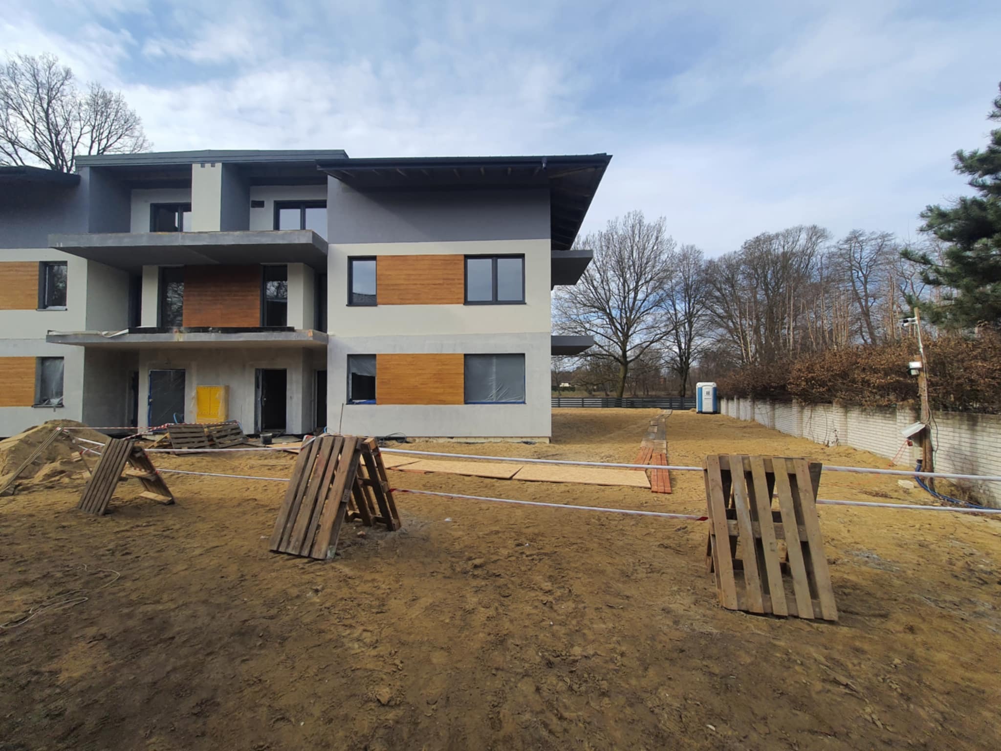 Wielopiętrowy budynek mieszkalny w trakcie budowy, z widoczną częścią elewacji w kolorach szarości i drewna, zabezpieczony na zewnątrz drewnianymi paletami i taśmą ostrzegawczą, z toaletą przenośną...