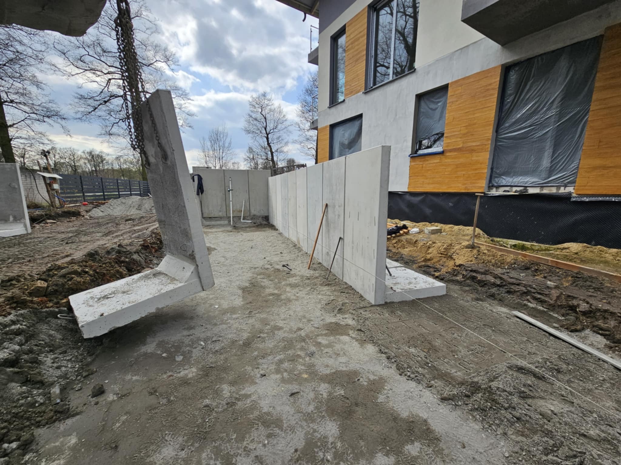 Montaż prefabrykowanych betonowych elementów konstrukcyjnych fundamentów przy nowo budowanym domu z częściowo wykończoną elewacją.