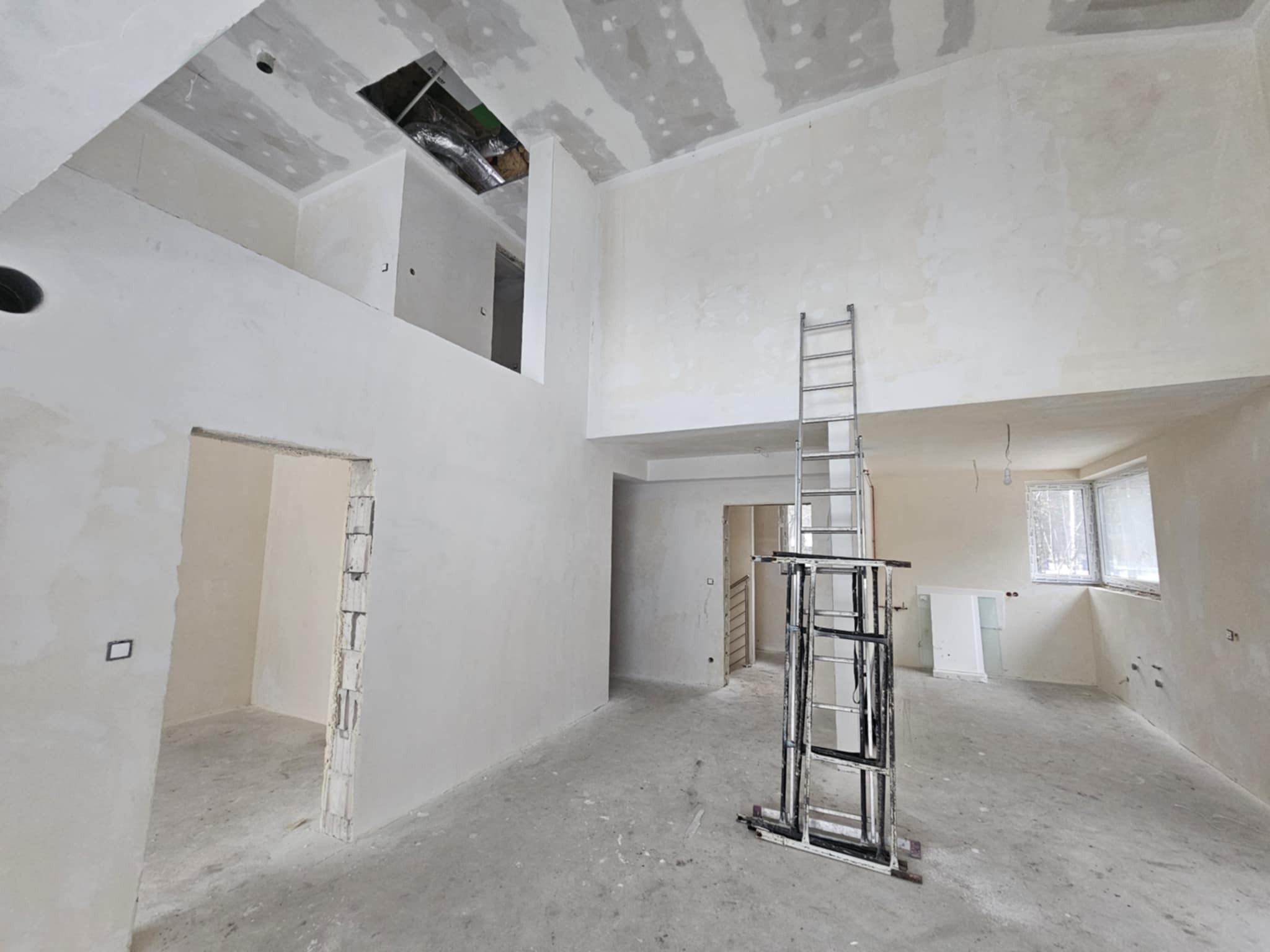 Wnętrze w stanie deweloperskim z widoczną antresolą, ścianami przygotowanymi do malowania, odsłoniętą instalacją w suficie i rozstawionym rusztowaniem.