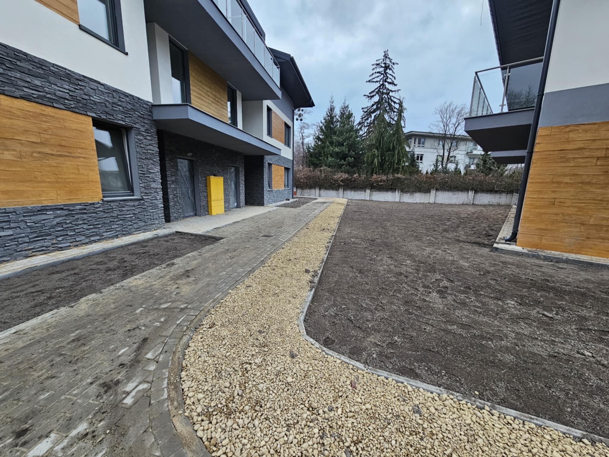 Dwukondygnacyjny budynek z elewacją wykończoną kamieniem, drewnem i białym tynkiem, widok z zewnątrz na świeżo przygotowany teren z kostką brukową i wysypanym drobnym kamieniem.