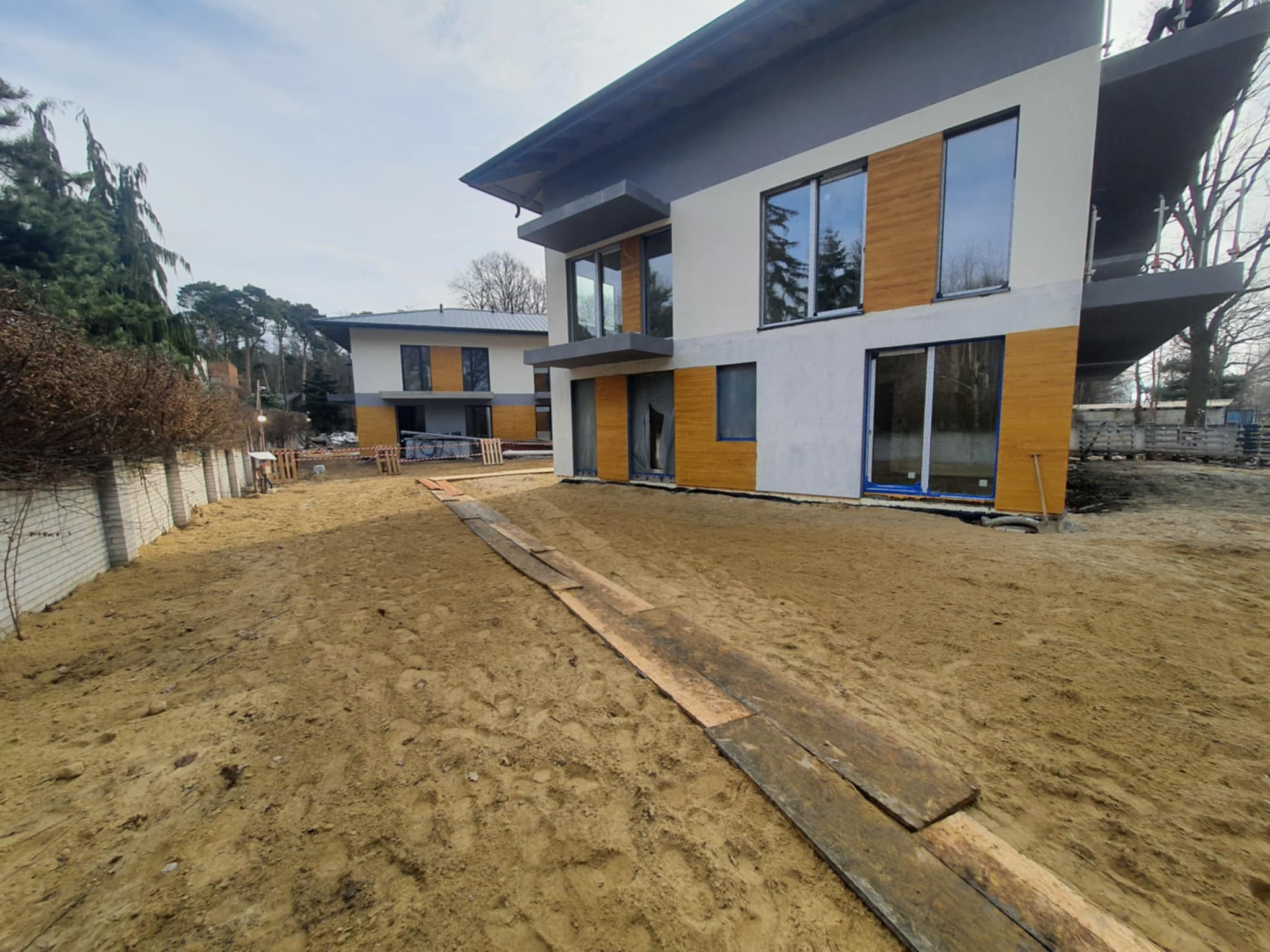 Dwa nowoczesne, dwukondygnacyjne domy murowane w trakcie budowy, z częściowo wykończoną elewacją w kolorze białym, szarym i z panelami drewnopodobnymi, widoczne tymczasowe chodniki z desek...