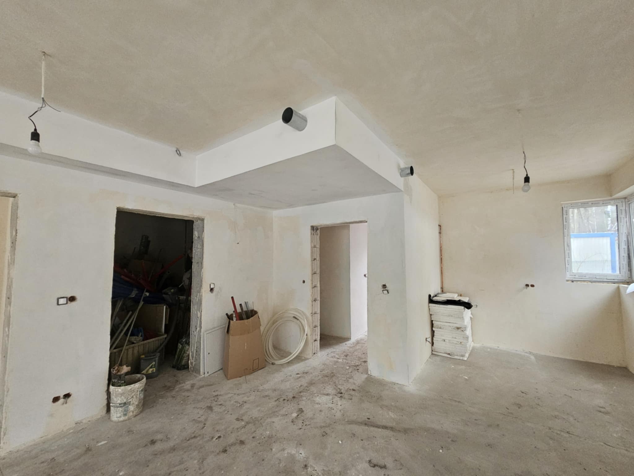 Surowy stan wnętrza domu w trakcie prac wykończeniowych: ściany po tynkowaniu, widoczne instalacje, prowizoryczne oświetlenie, podłoga wylewka, materiały budowlane w pomieszczeniu.