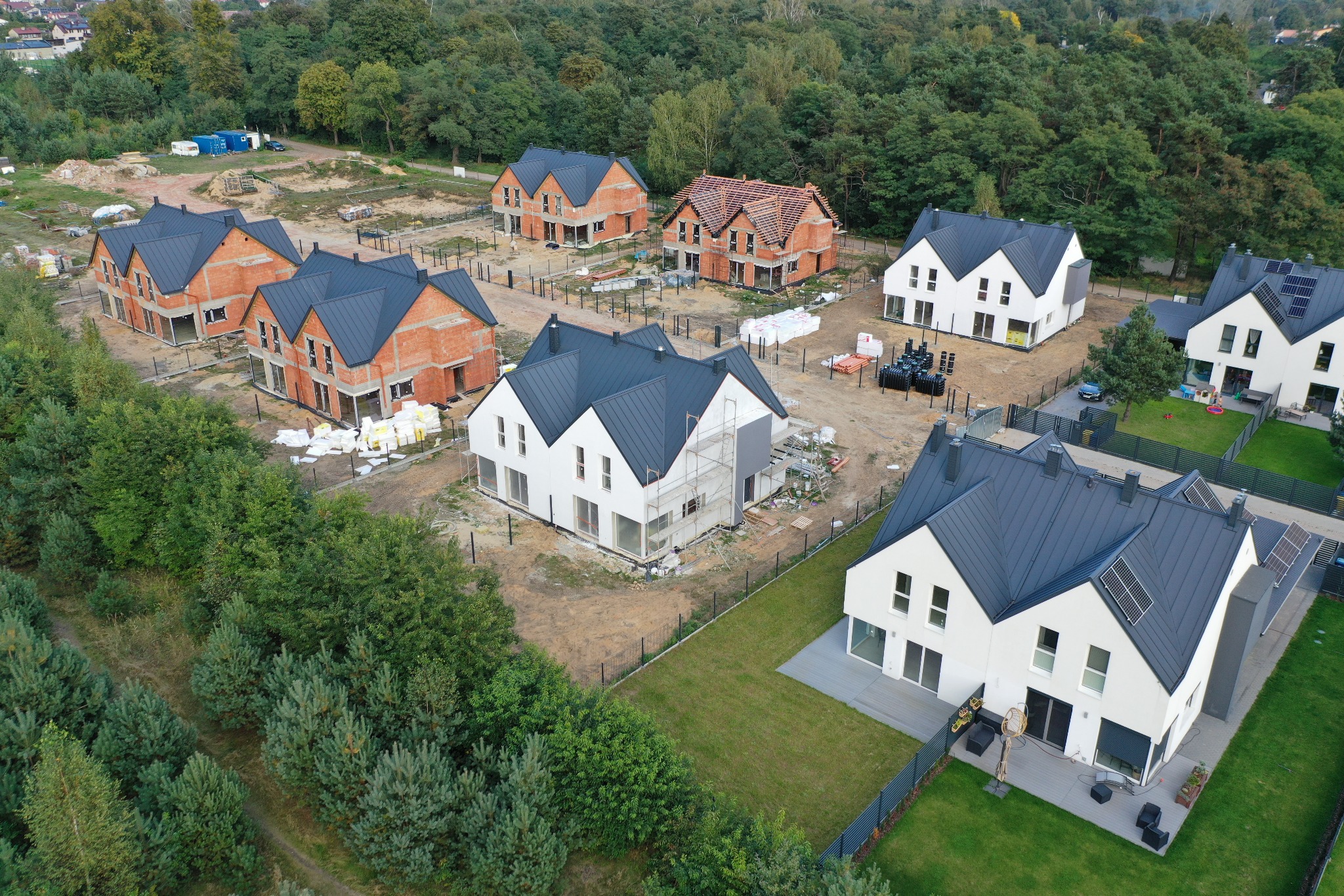 Widok z lotu ptaka na osiedle domów jednorodzinnych w różnym stadium budowy, od surowej cegły po domy z panelami słonecznymi i zagospodarowanym terenem.