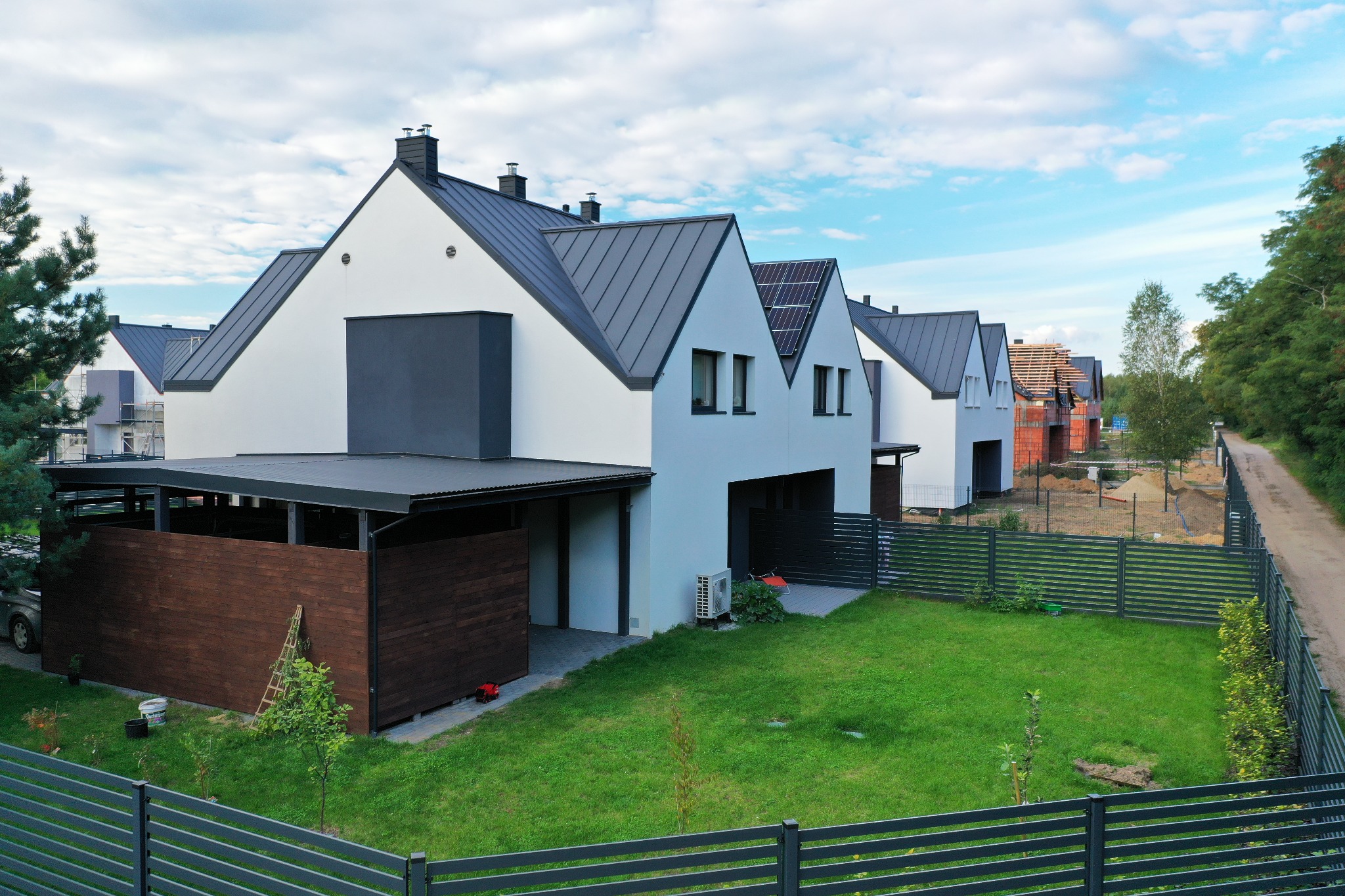 Szereg nowoczesnych domów jednorodzinnych z białymi ścianami, grafitowymi dachami i panelami słonecznymi, widok z zewnątrz na ogrodzony teren z zielonym trawnikiem, w tle dom w trakcie budowy...