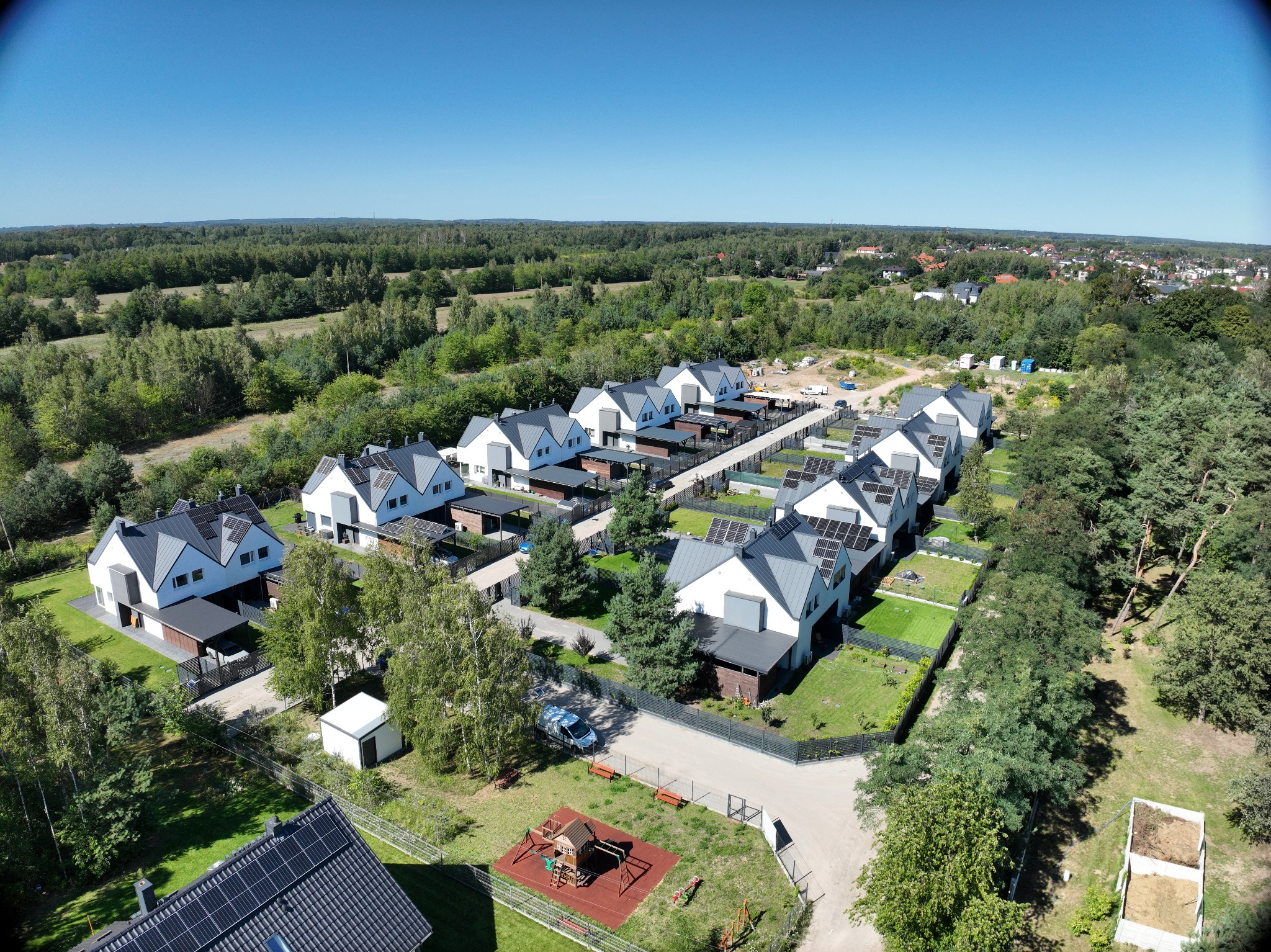 Osiedle nowoczesnych domów jednorodzinnych z panelami słonecznymi na dachach, widok z lotu ptaka na tle lasu i błękitnego nieba.