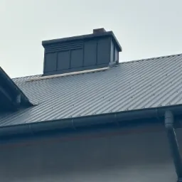Szary dach z blachy trapezowej z kominem wentylacyjnym, widoczny system rynnowy z bliska.
