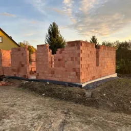 Wznoszone mury parterowego domu z pustaków ceramicznych, widoczna izolacja przeciwwilgociowa fundamentów i fragment istniejącego budynku w tle.