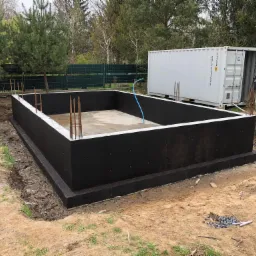 Zabezpieczony fundament pod budynek, widoczny w wykopie, z wystającymi prętami zbrojeniowymi, obok kontener budowlany i narzędzia.