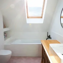 Jasna łazienka na poddaszu z różową heksagonalną podłogą, białą wanną obudowaną płytkami, drewnianą szafką z nablatową umywalką i oknem dachowym wpuszczającym silne światło.
