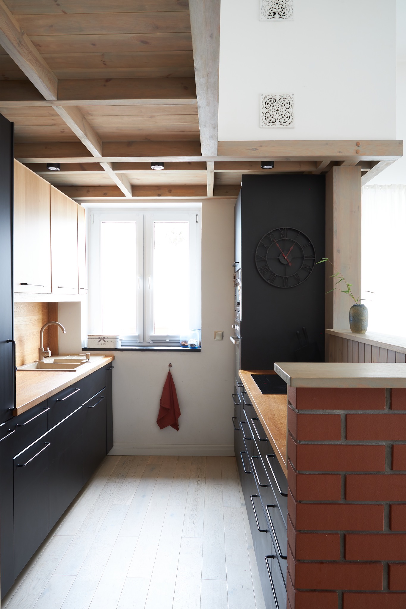 Wnętrze kuchni z drewnianym sufitem, czarnymi szafkami i ceglaną ścianką działową, widok na okno z suszącą się ścierką.