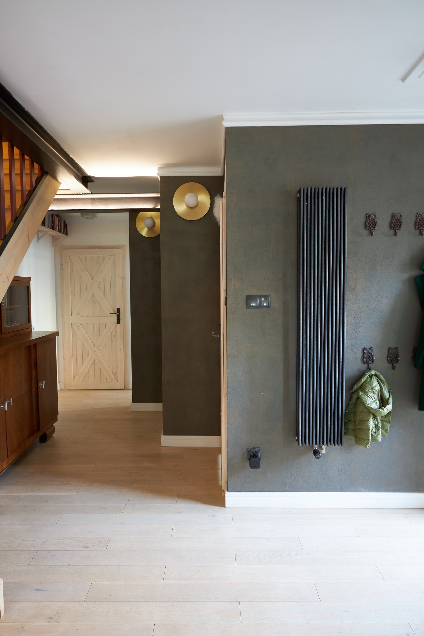 Wnętrze przedpokoju w Poznaniu z drewnianą podłogą, szarą ścianą z grzejnikiem panelowym, złotymi lampami ściennymi i wieszakami w kształcie sów.
