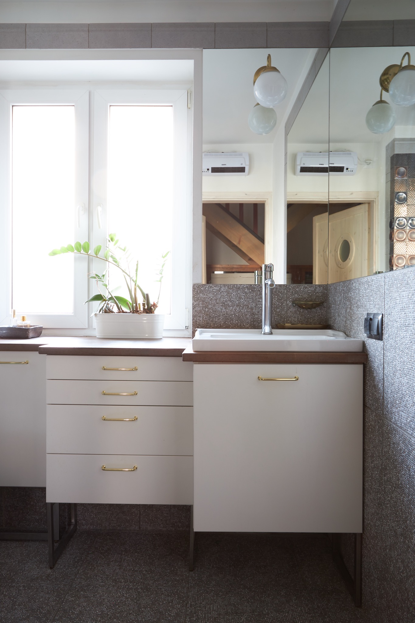 Nowoczesna łazienka z umywalką nablatową, szafką z szufladami na metalowych nóżkach i dużym lustrem, na blacie doniczka z rośliną, widoczne schody w odbiciu lustra.