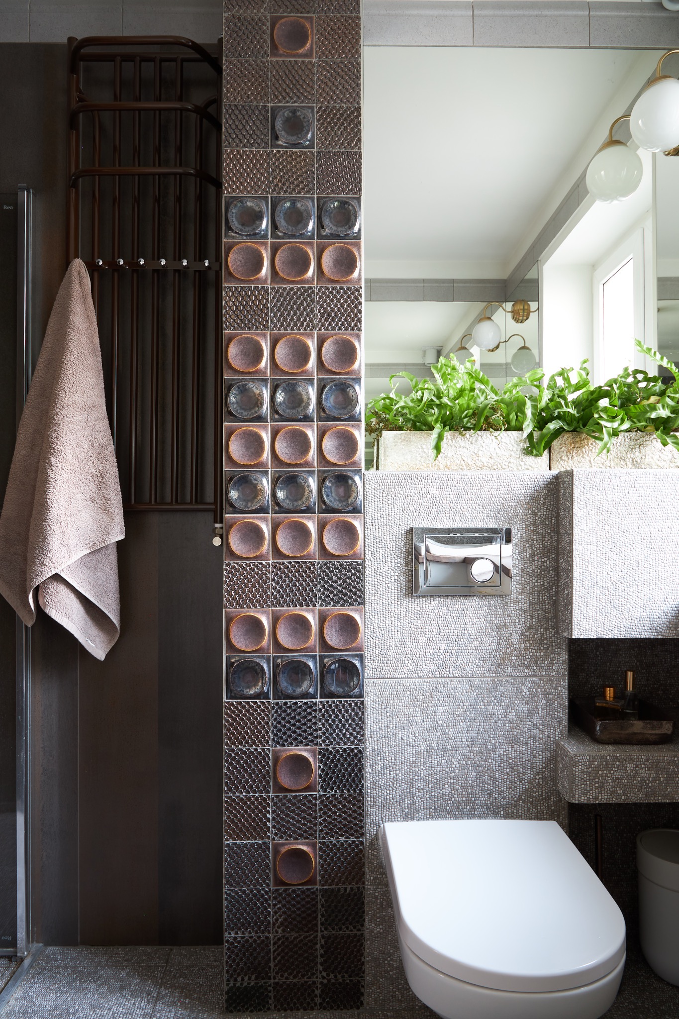 Nowoczesna łazienka z oryginalnymi, brązowo-szarymi płytkami z okrągłym motywem, wiszącą toaletą, roślinami w donicach i grzejnikiem drabinkowym z ręcznikiem.